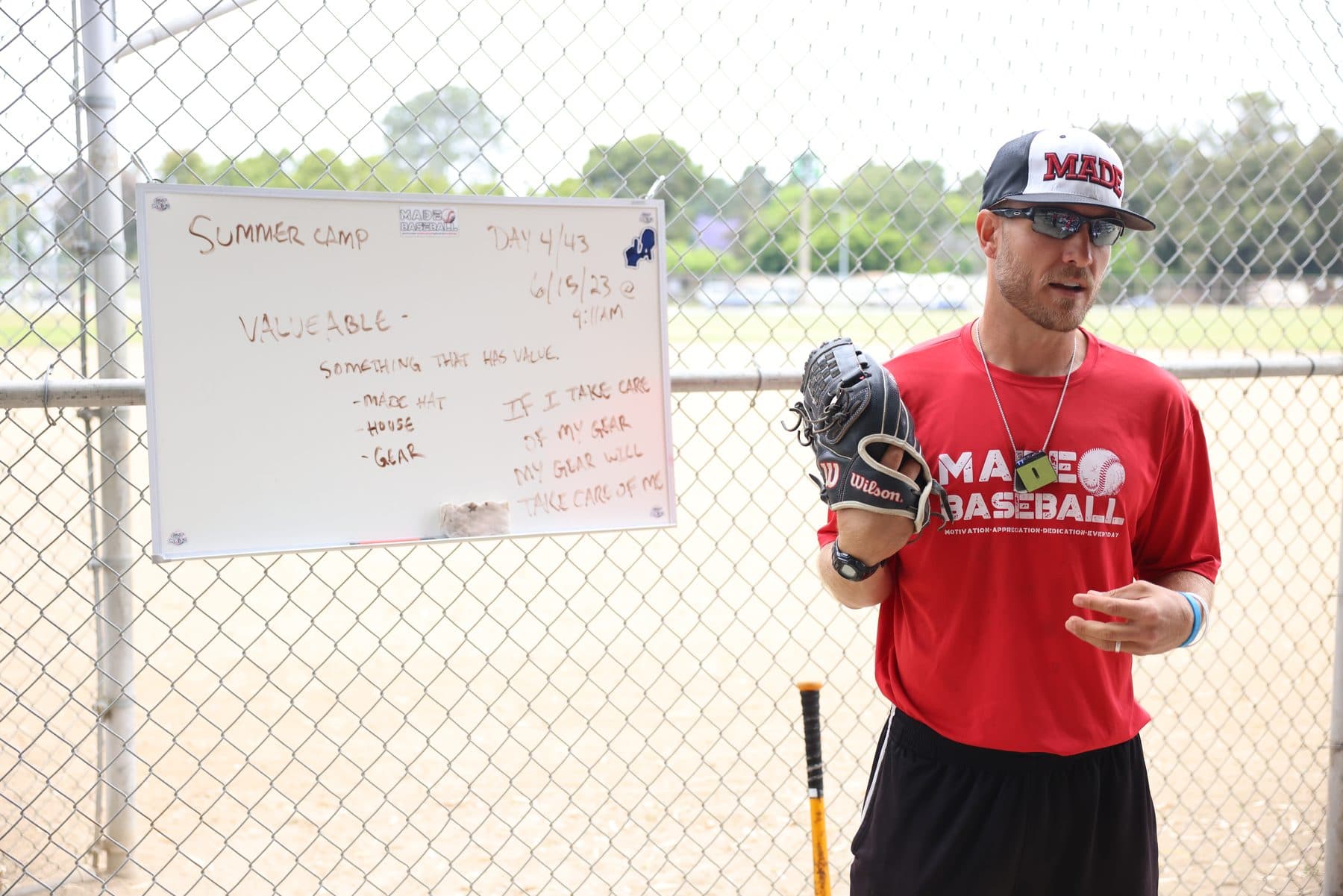 Coach Isaac Hess in a red MADE Baseball shirt and cap, glove in hand, talking beside a whiteboard