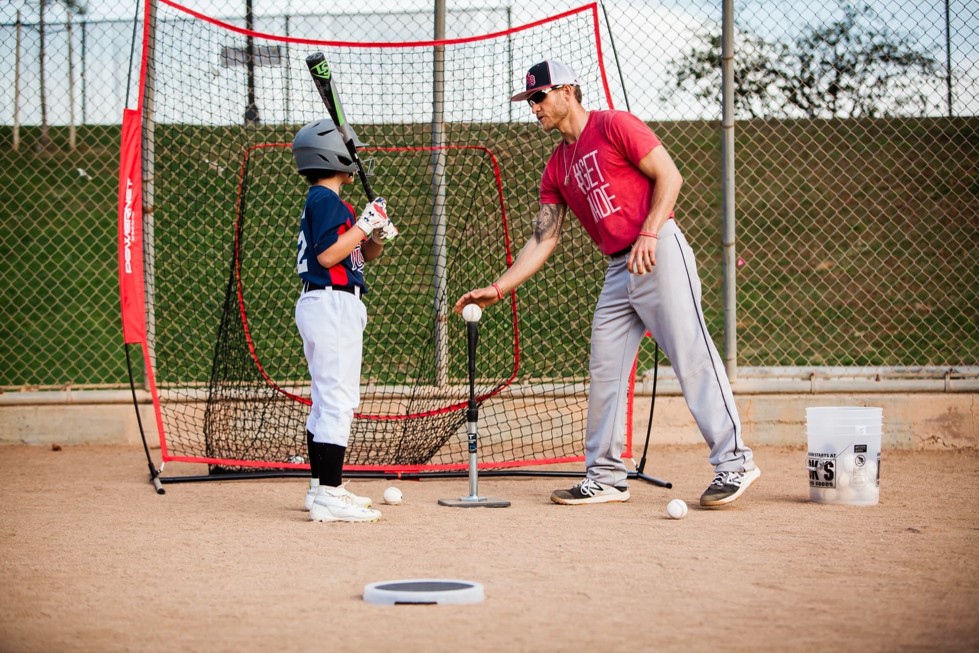 Coach Isaac Hess in a red shirt placing a ball on the tee while a young hitter in a helmet waits in his stance, ready to swing